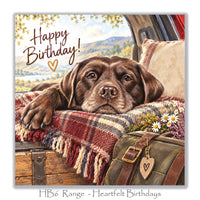 Charming dog birthday greeting card with a Chocolate Labrador, cosy red tartan blanket, rustic suitcase, wildflowers and peaceful countryside lake.