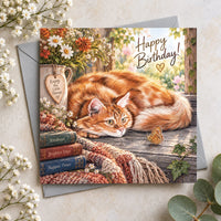 Cat birthday card featuring a ginger cat lying by a window with flowers, books, butterfly and Happy Birthday message.