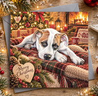 Staffy puppy Christmas card featuring a cute white and brown Staffordshire Bull Terrier resting on a tartan blanket beside a Christmas tree and fireplace.