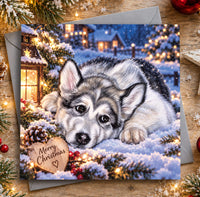 Alaskan Malamute Christmas card featuring an adorable fluffy puppy resting in the snow beside a glowing lantern and Merry Christmas wooden heart.