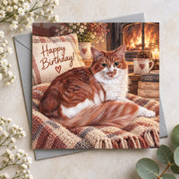 Ginger cat birthday card featuring a beautiful fluffy ginger and white cat resting beside a glowing fireplace with Happy Birthday lettering.