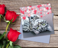 Cute tabby kitten greeting card featuring a fluffy kitten with green eyes lying on his back beneath heart-shaped bunting.