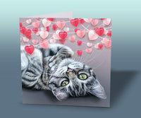 Unique kitten art card with a green-eyed tabby kitten lying comfortably under decorative hearts, printed in the UK.