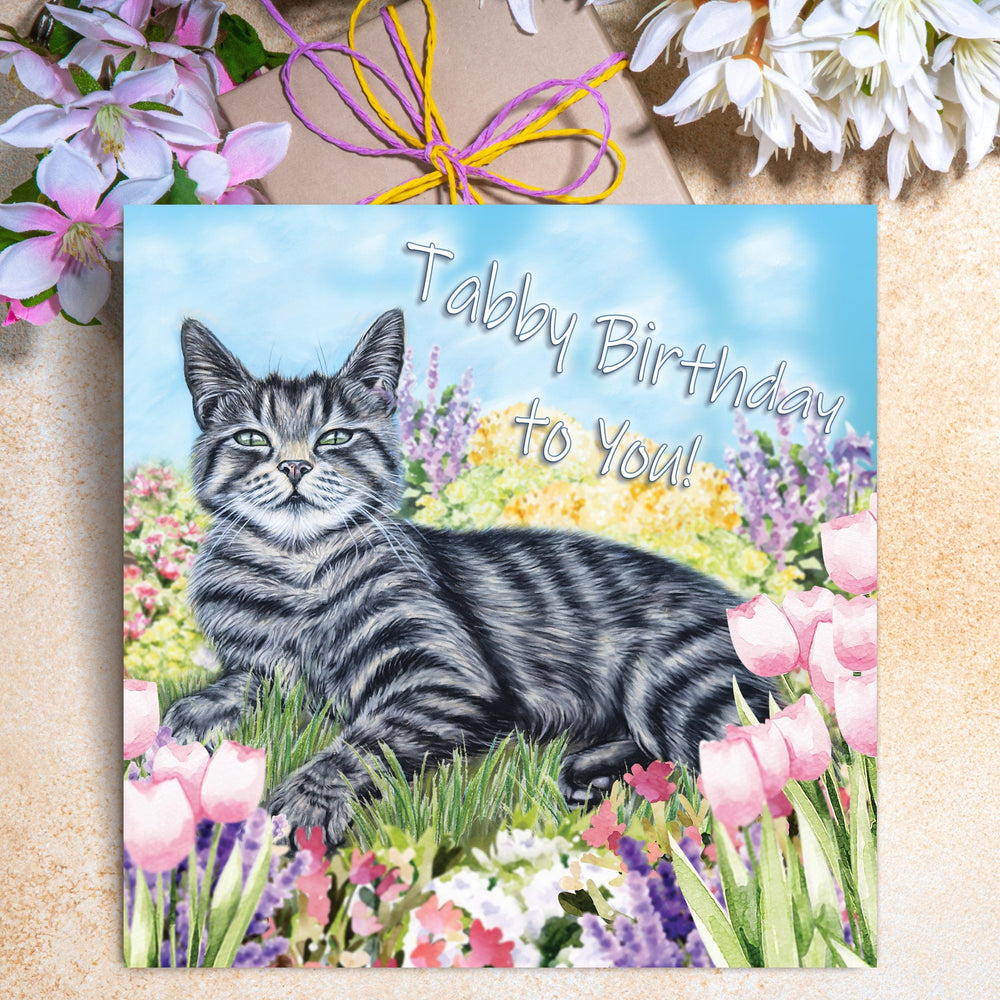 Tabby cat birthday greeting card featuring a proud tabby lounging under a blue sky surrounded by colourful flowers, with the words Tabby Birthday to You.